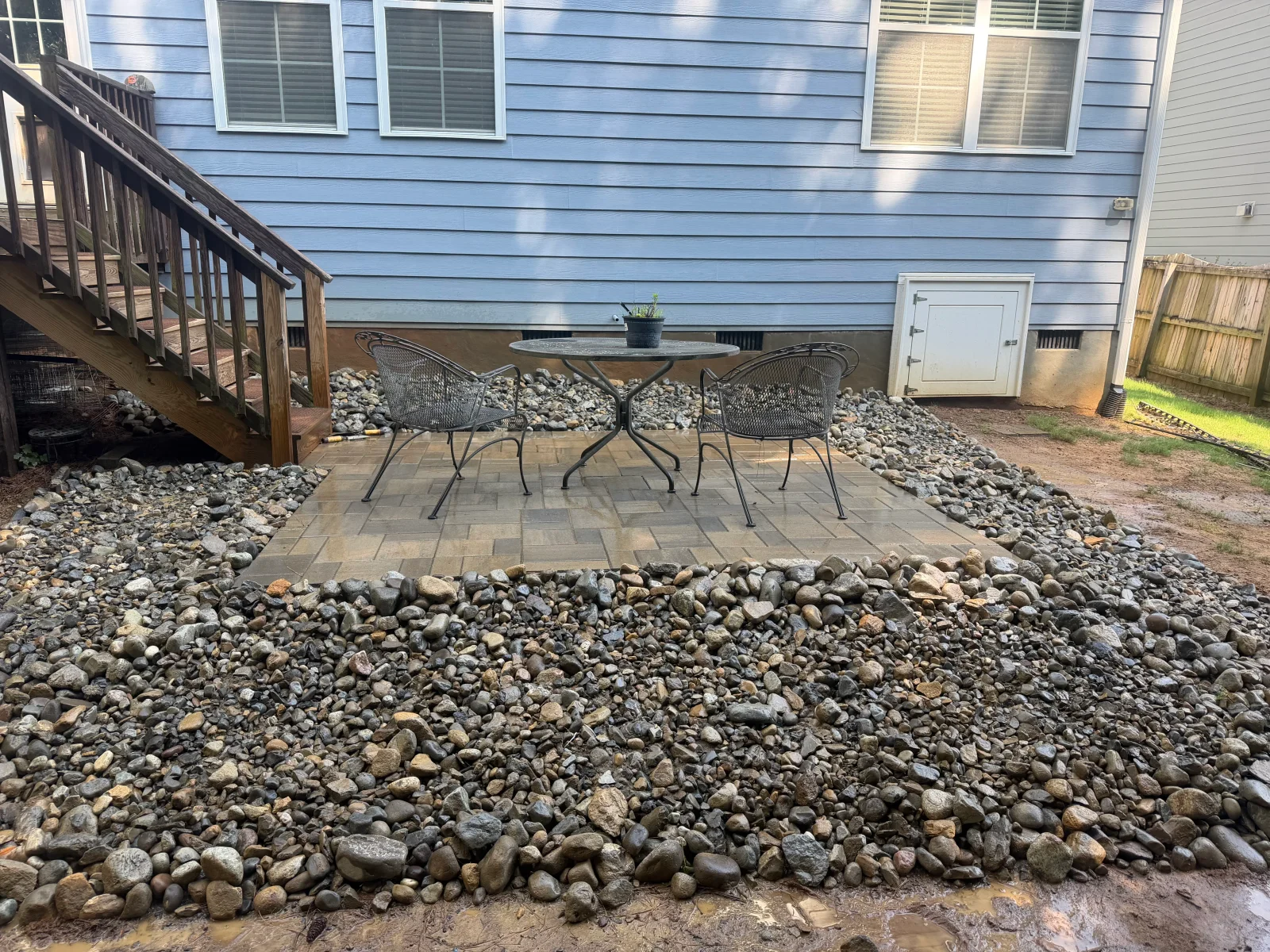 Rectangular paver patio with metal seating, bordered by river rock and stone edging against blue house