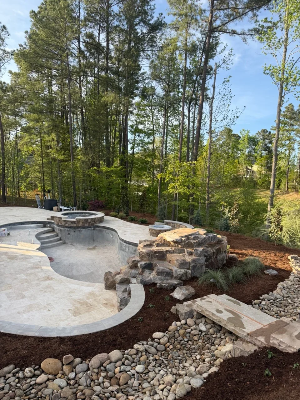 Concrete pool deck with curved design, stone fire pit, river rock borders, and wooded backdrop