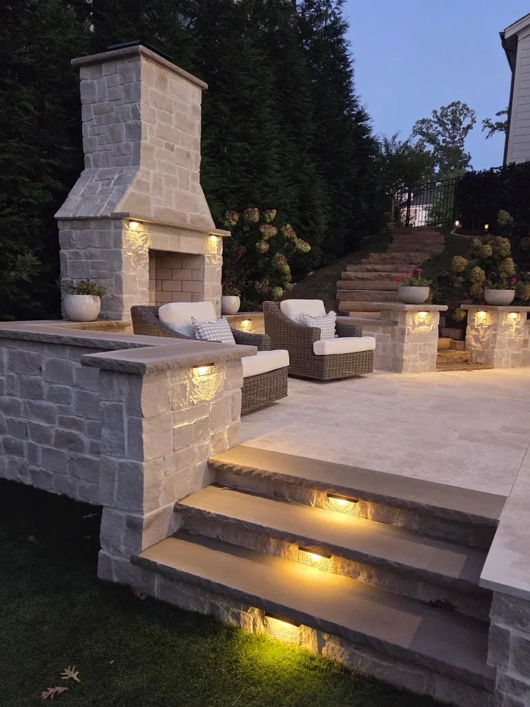 Illuminated stone fireplace and patio with seating, step lights, and landscape lighting at dusk
