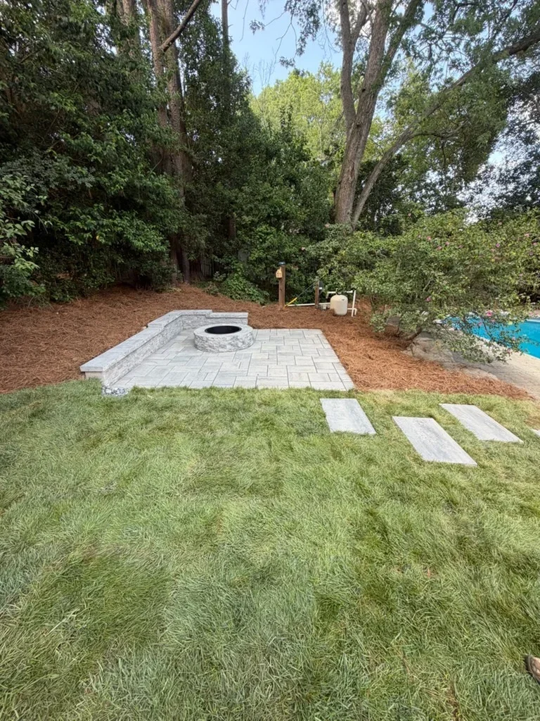 Gray paver patio with circular fire pit, mulch beds, and stepping stones in wooded yard