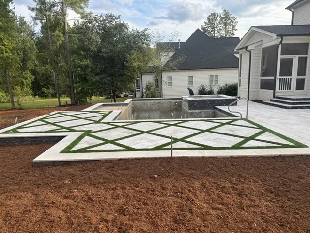 Concrete patio with geometric green lawn pattern beside in-ground pool and home